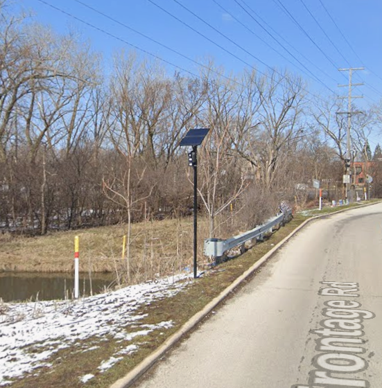 A Flock Safety camera in a black housing mounted on a pole beside a road, with a small solar panel above it.